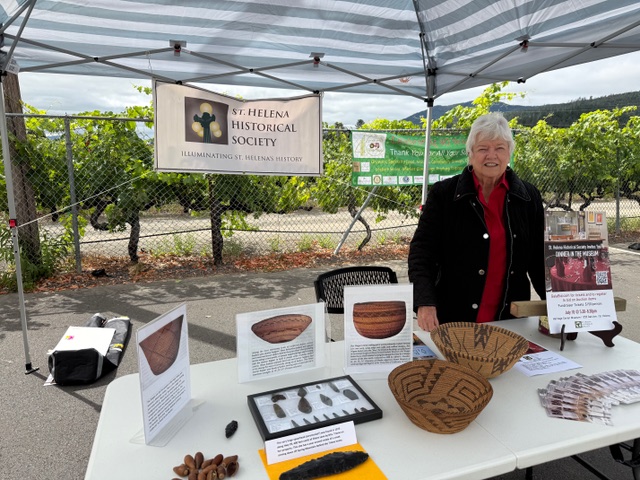 Two large coiled Wappo baskets and stone artifacts on the St. Helena Historical Society information table, with the Society banner and a vineyard behind.