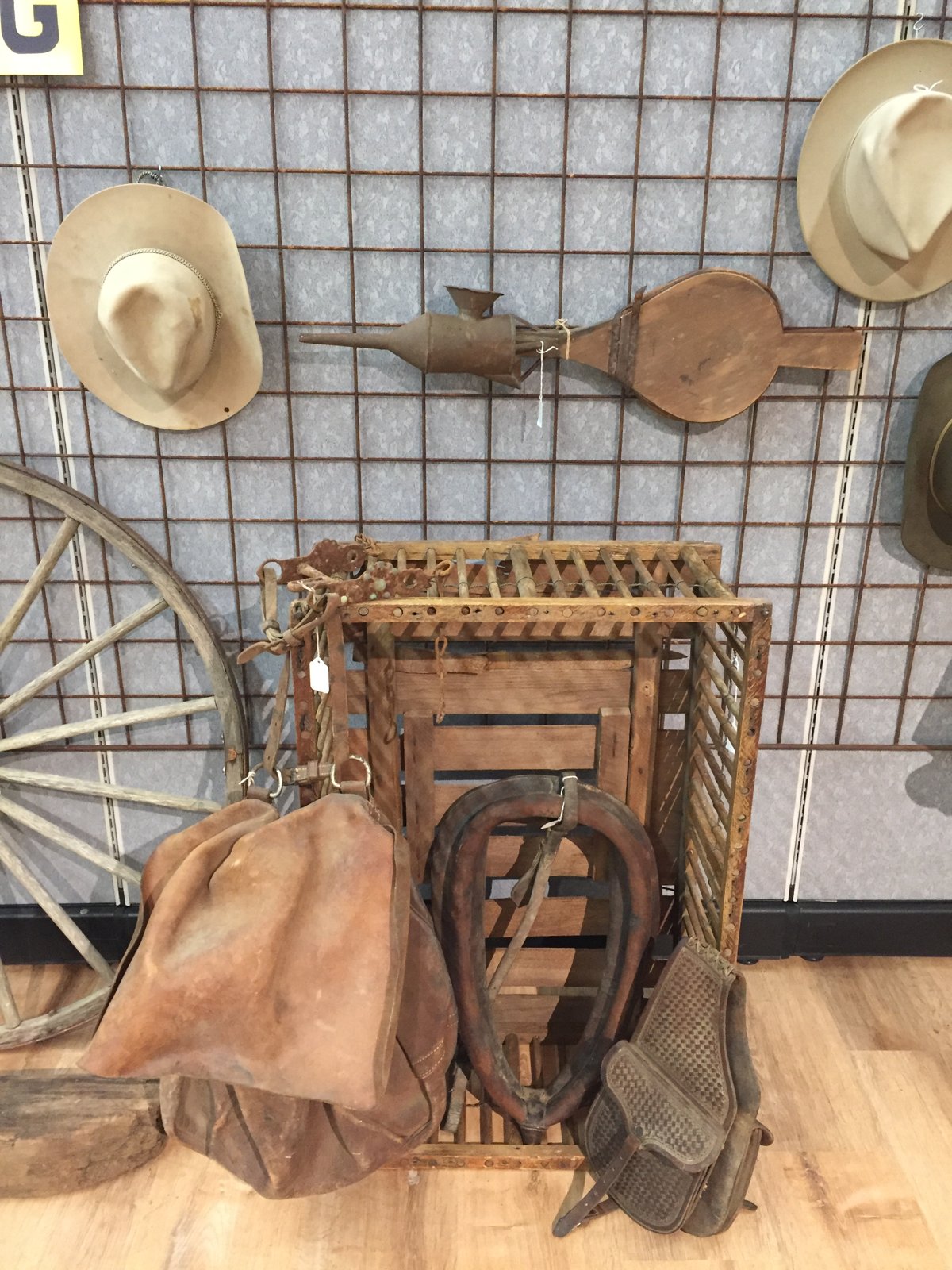 Antique saddles, horse collar, hats, bellows and a wagon wheel displayed on a museum wall.