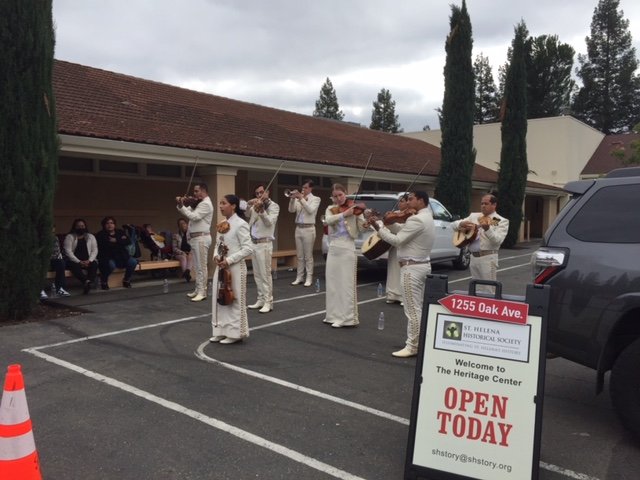 A mariachi band in white charro suits plays violins, guitars and trumpets in the lot outside the Heritage Center, beside a Heritage Center Open Today sign.