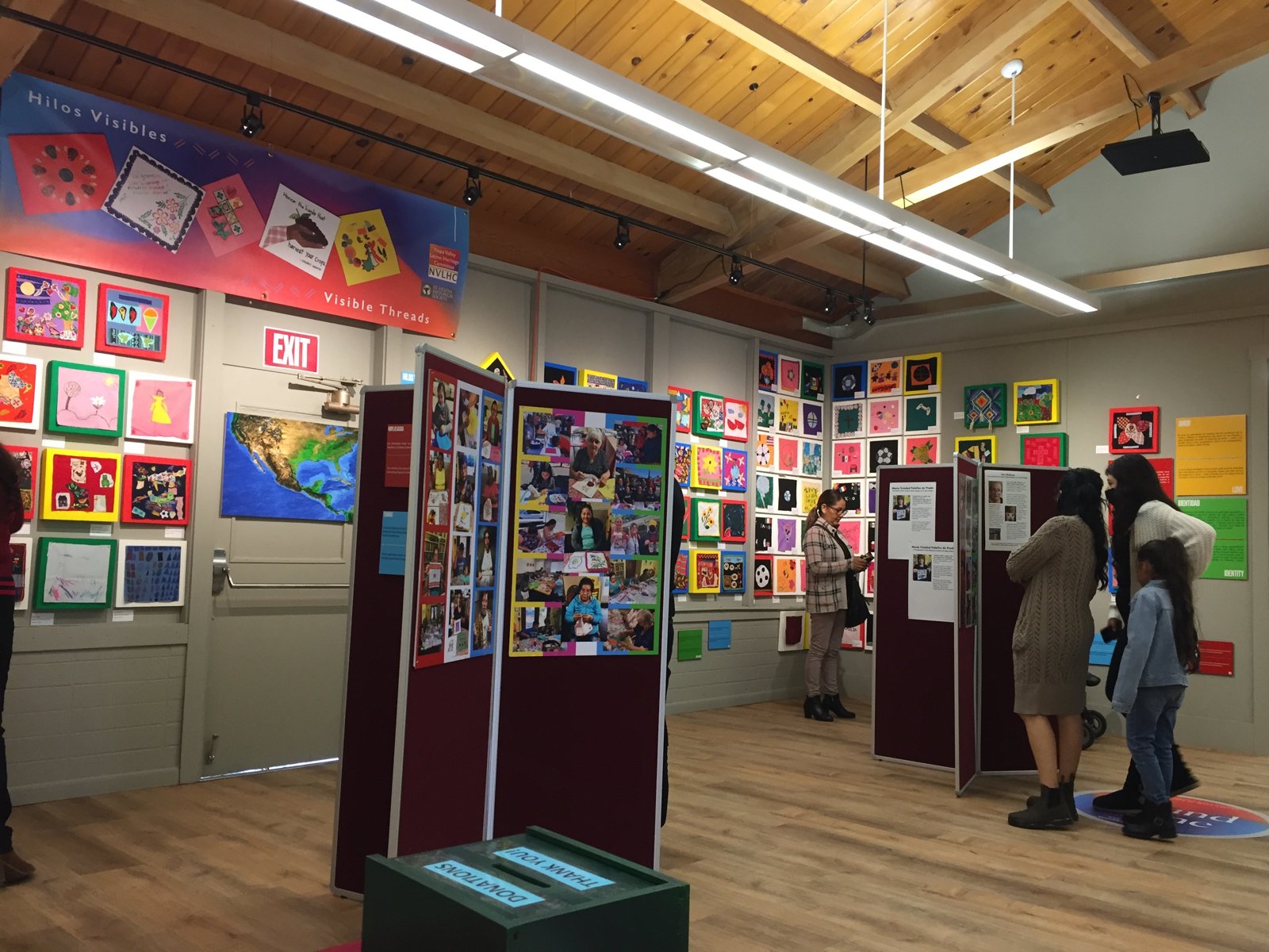 Visitors viewing colorful textile and mixed-media artworks at the Hilos Visibles / Visible Threads exhibit, with a donations box in the foreground.