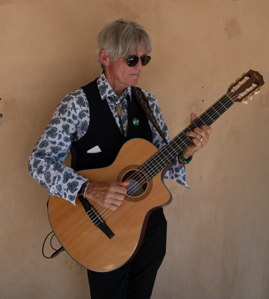 A guitarist in a patterned shirt and vest plays a classical guitar.